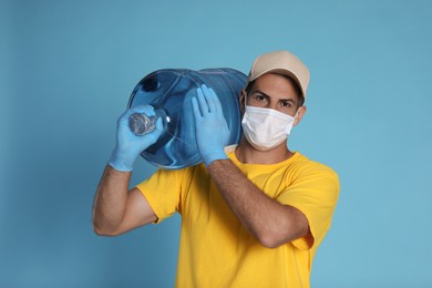 Photo of Courier in medical mask holding bottle for water cooler on light blue background. Delivery during coronavirus quarantine