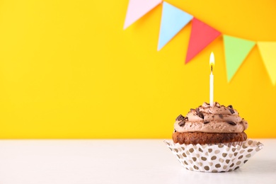 Photo of Chocolate cupcake with burning candle on white table against yellow background. Space for text