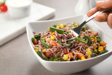 Photo of Woman eating tasty brown rice with vegetables at grey table, closeup