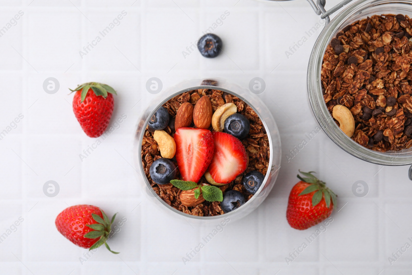 Photo of Tasty granola with berries, nuts and mint in glass on white tiled table, flat lay