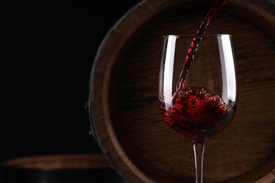 Photo of Pouring red wine into glass near wooden barrel against black background, closeup. Space for text