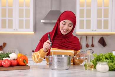 Muslim woman making delicious soup with vegetables at white table in kitchen