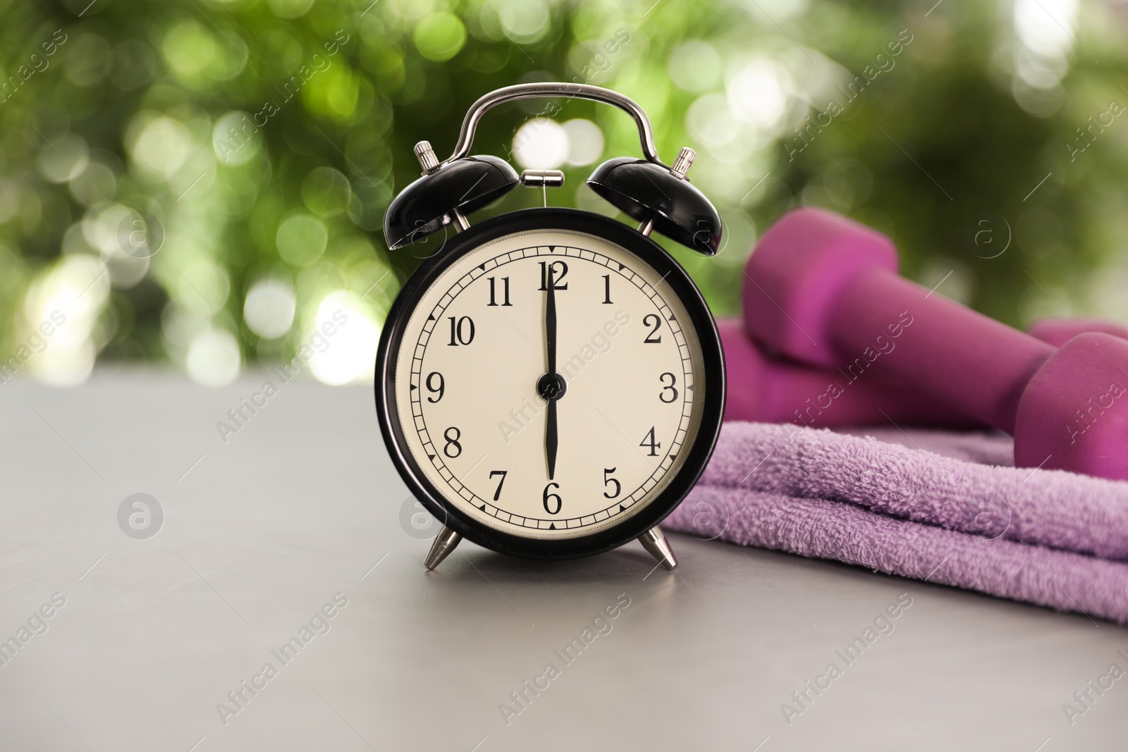 Photo of Alarm clock, towel and dumbbells on grey table against blurred background. Morning exercise