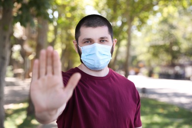 Photo of Man in protective face mask showing stop gesture outdoors. Prevent spreading of coronavirus