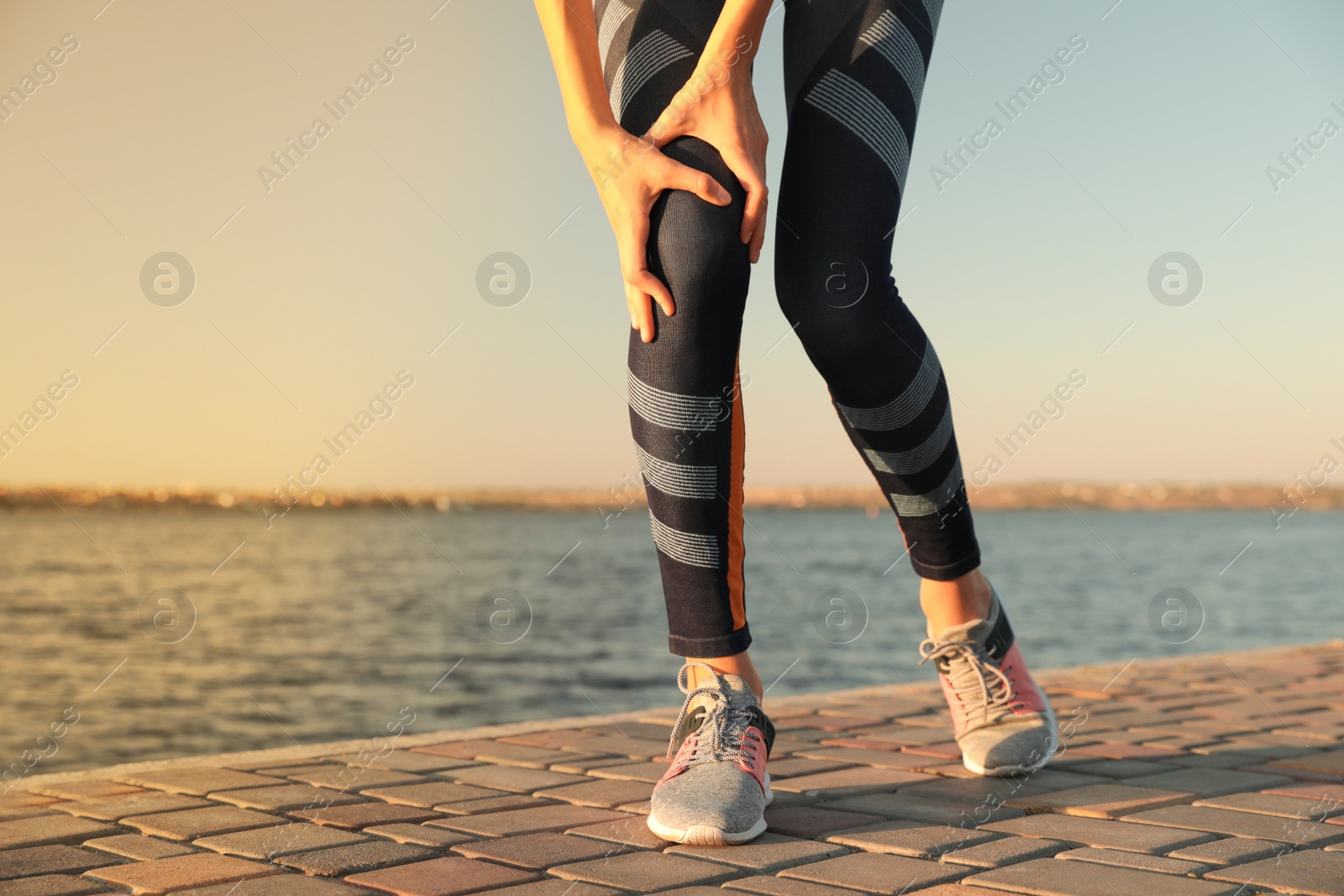 Photo of Young woman in sportswear having knee problems near river at sunset, closeup