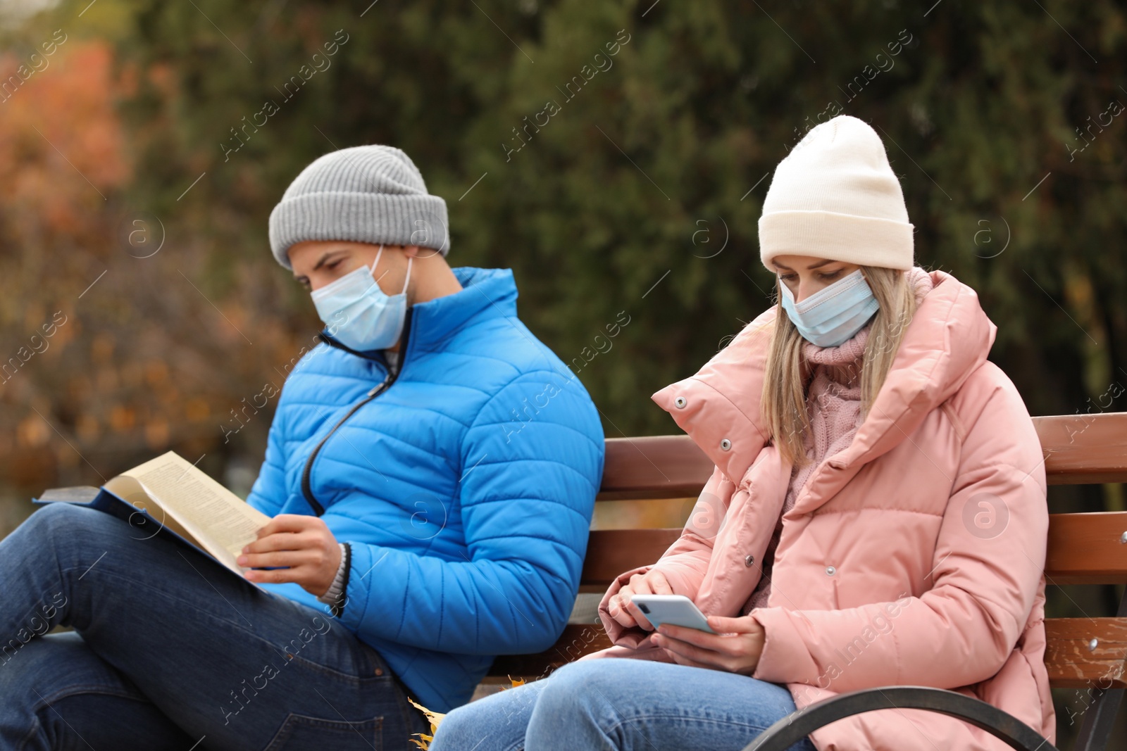 Photo of People in medical masks keeping distance while sitting on bench outdoors. Protective measures during coronavirus quarantine