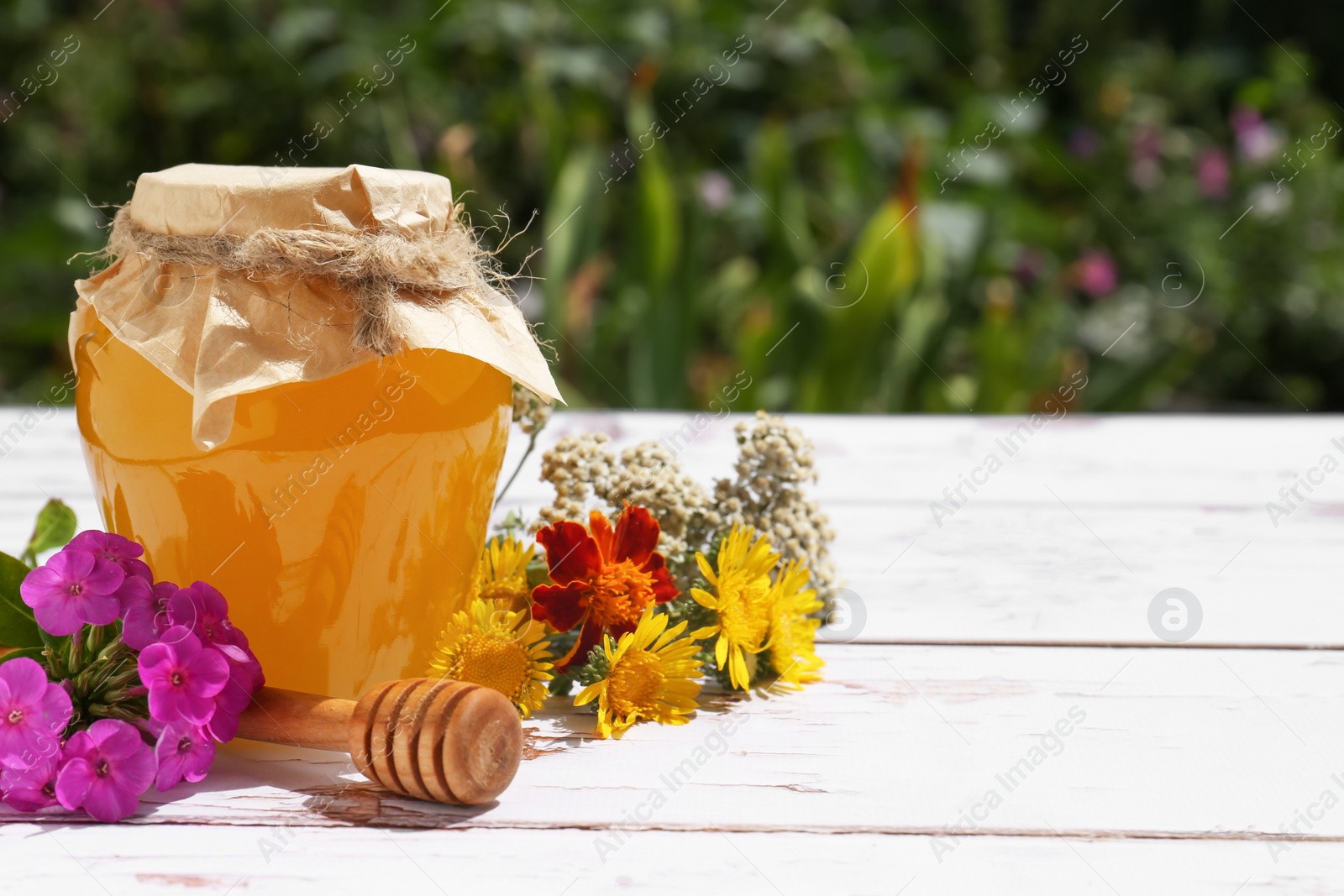 Photo of Glass jar of honey, dipper and different flowers on white wooden table in garden. Space for text