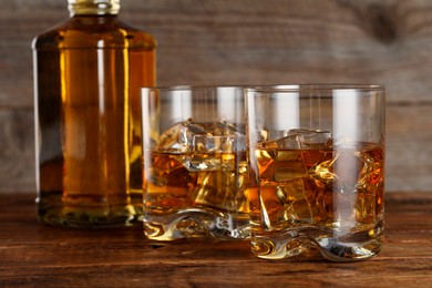 Photo of Whiskey with ice cubes in glasses and bottle on wooden table, closeup