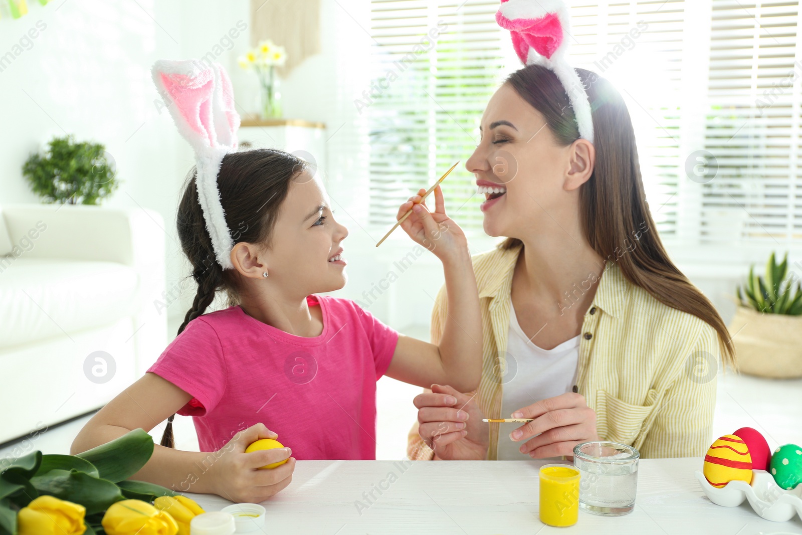 Photo of Happy mother and daughter with bunny ears headbands having fun while painting Easter egg at home