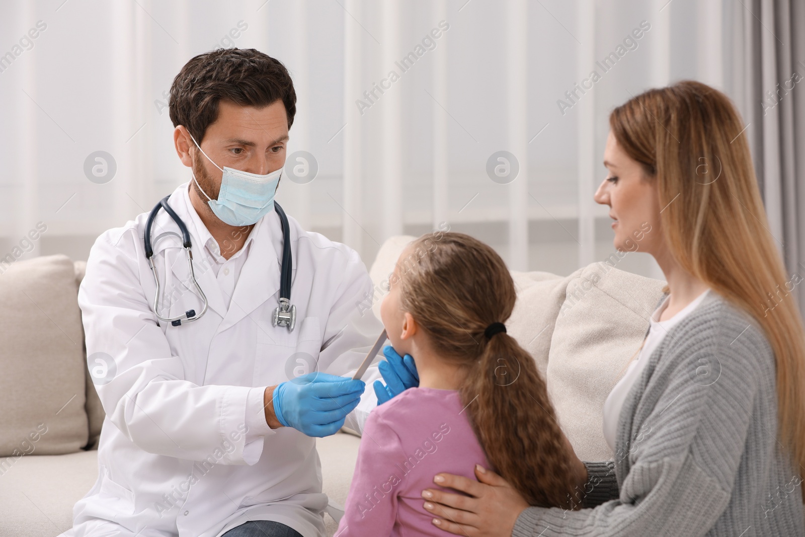 Photo of Doctor in medical mask examining girl`s oral cavity with tongue depressor near her mother indoors