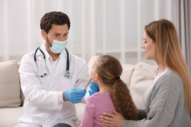 Photo of Doctor in medical mask examining girl`s oral cavity with tongue depressor near her mother indoors