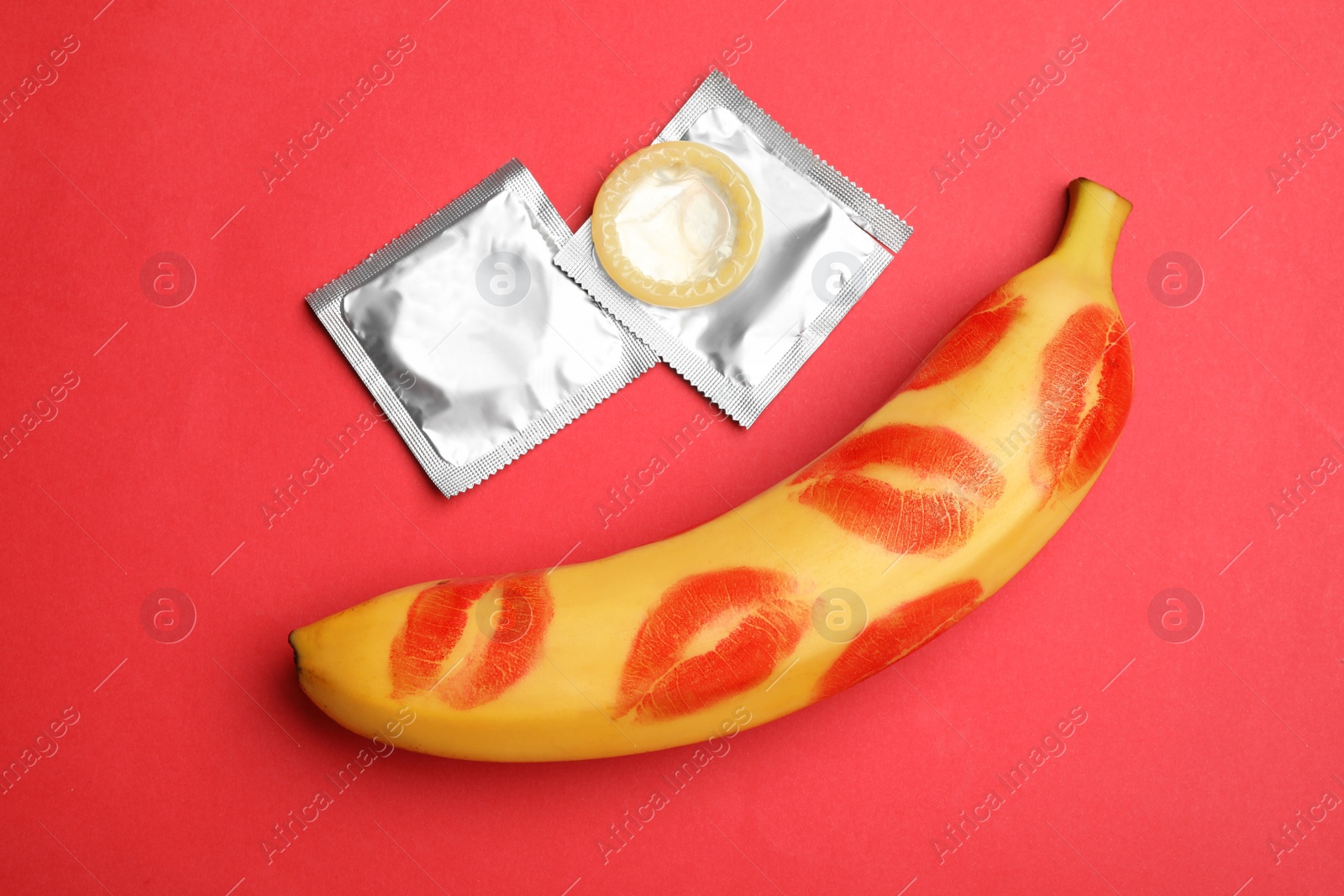 Photo of Condoms and banana with lipstick kiss marks on red background, flat lay. Safe sex