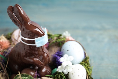 Photo of Chocolate bunny with protective mask, eggs, wreath and space for text on light blue wooden table, closeup. Easter holiday during COVID-19 quarantine