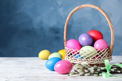 Photo of Wicker basket with bright painted Easter eggs and pussy willows on white wooden table against blue background. Space for text
