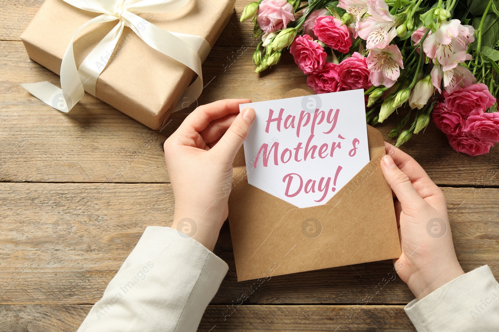 Image of Happy Mother's Day. Woman taking greeting card out of envelope, closeup. Gift box and bouquet of beautiful flowers on wooden table