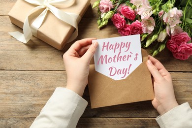 Happy Mother's Day. Woman taking greeting card out of envelope, closeup. Gift box and bouquet of beautiful flowers on wooden table