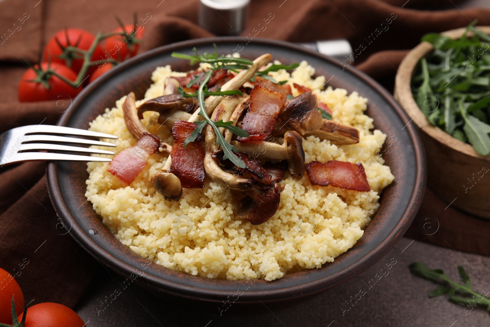 Photo of Bowl of tasty couscous with mushrooms, bacon and fork on brown table, closeup