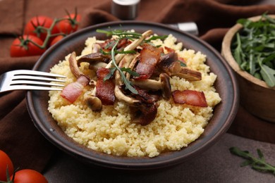 Photo of Bowl of tasty couscous with mushrooms, bacon and fork on brown table, closeup