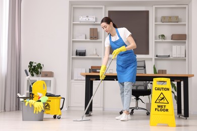 Cleaning service worker washing floor with mop. Bucket with supplies and wet floor sign in office