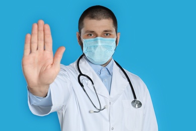 Photo of Doctor in protective mask showing stop gesture on light blue background. Prevent spreading of coronavirus