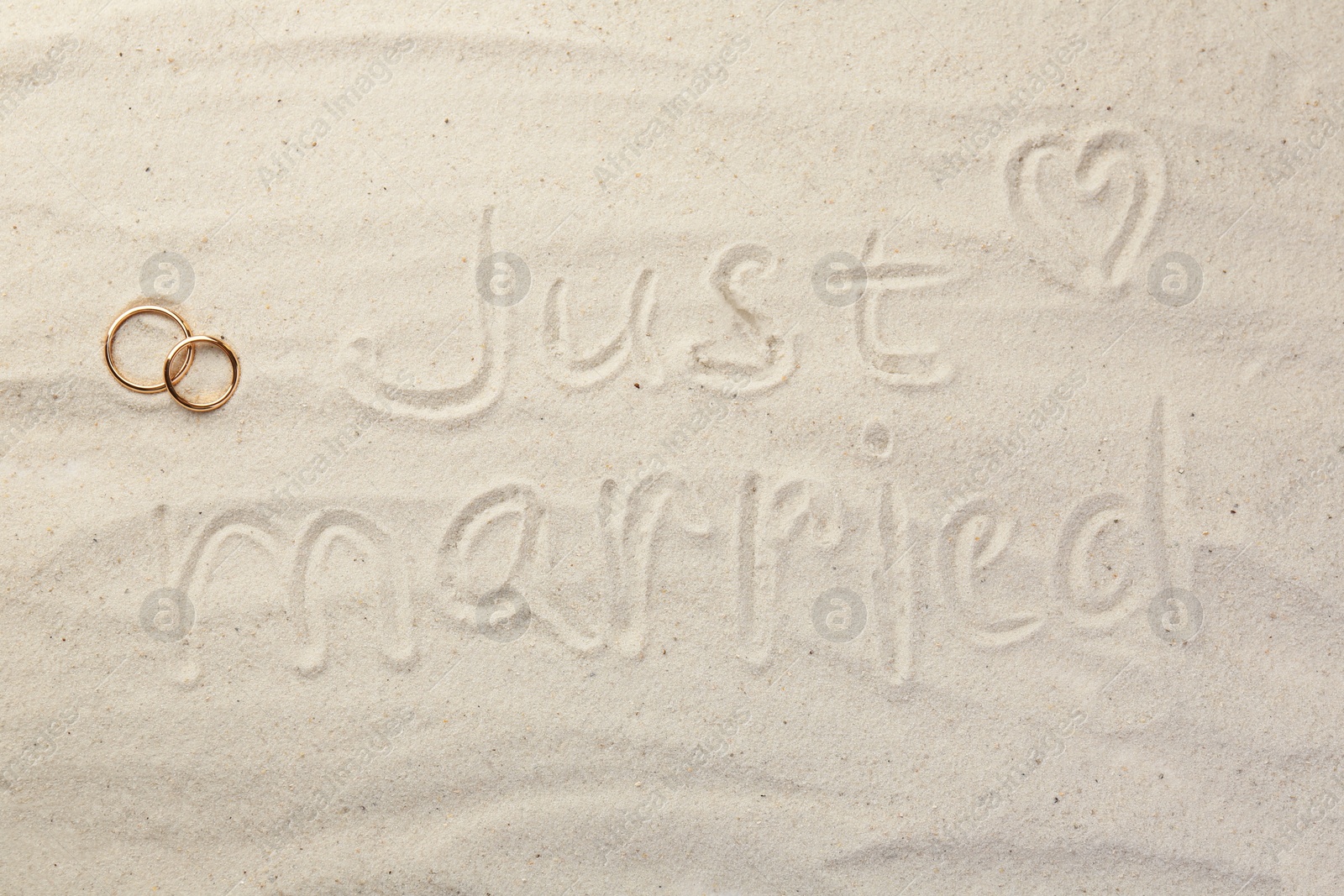 Photo of Honeymoon concept. Two golden rings and phrase Just married written on sand, top view