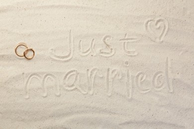 Photo of Honeymoon concept. Two golden rings and phrase Just married written on sand, top view