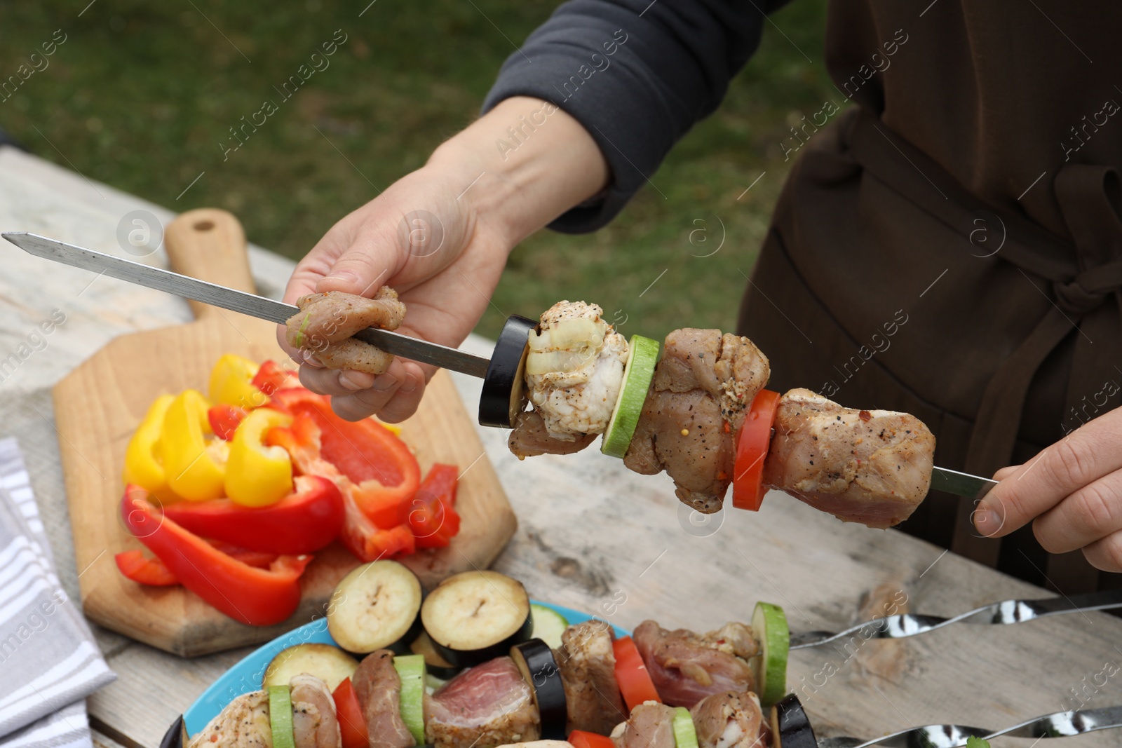 Photo of Woman stringing marinated meat and vegetables on skewer at wooden table outdoors, closeup