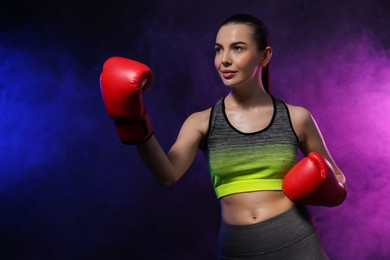 Photo of Portrait of beautiful woman wearing boxing gloves training in color lights and smoke on black background