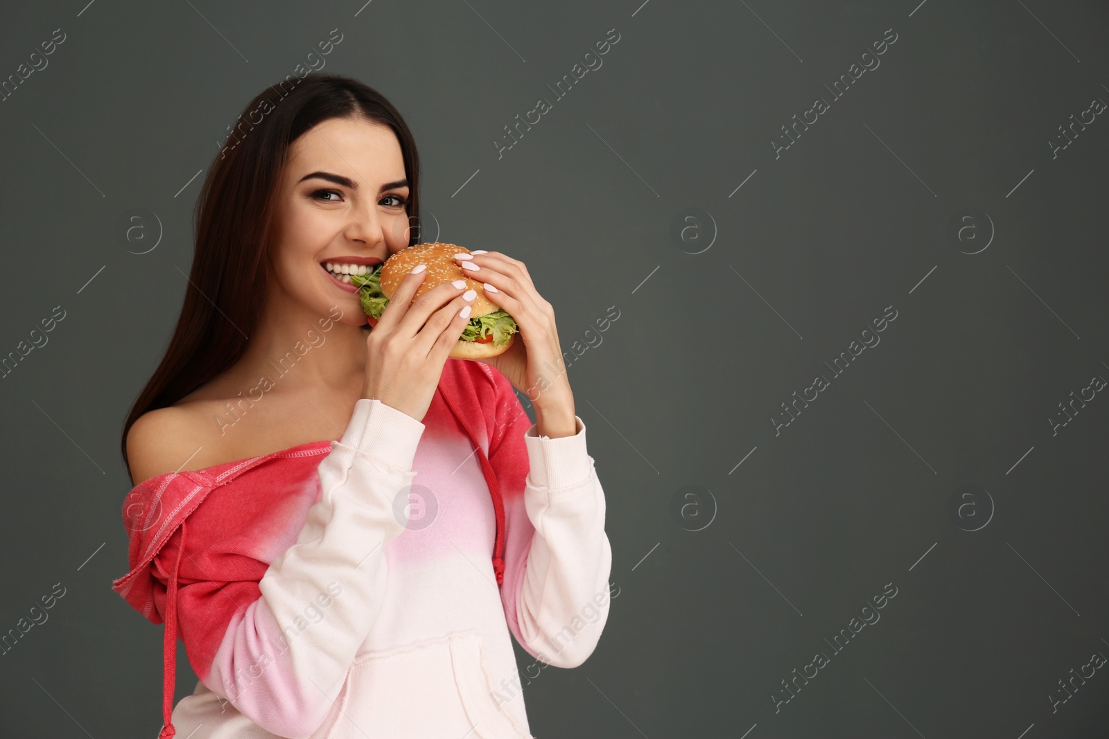 Photo of Young woman eating tasty burger on grey background. Space for text