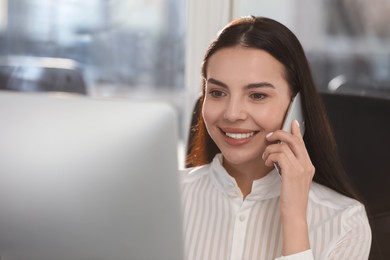 Happy woman using modern computer while talking on smartphone in office
