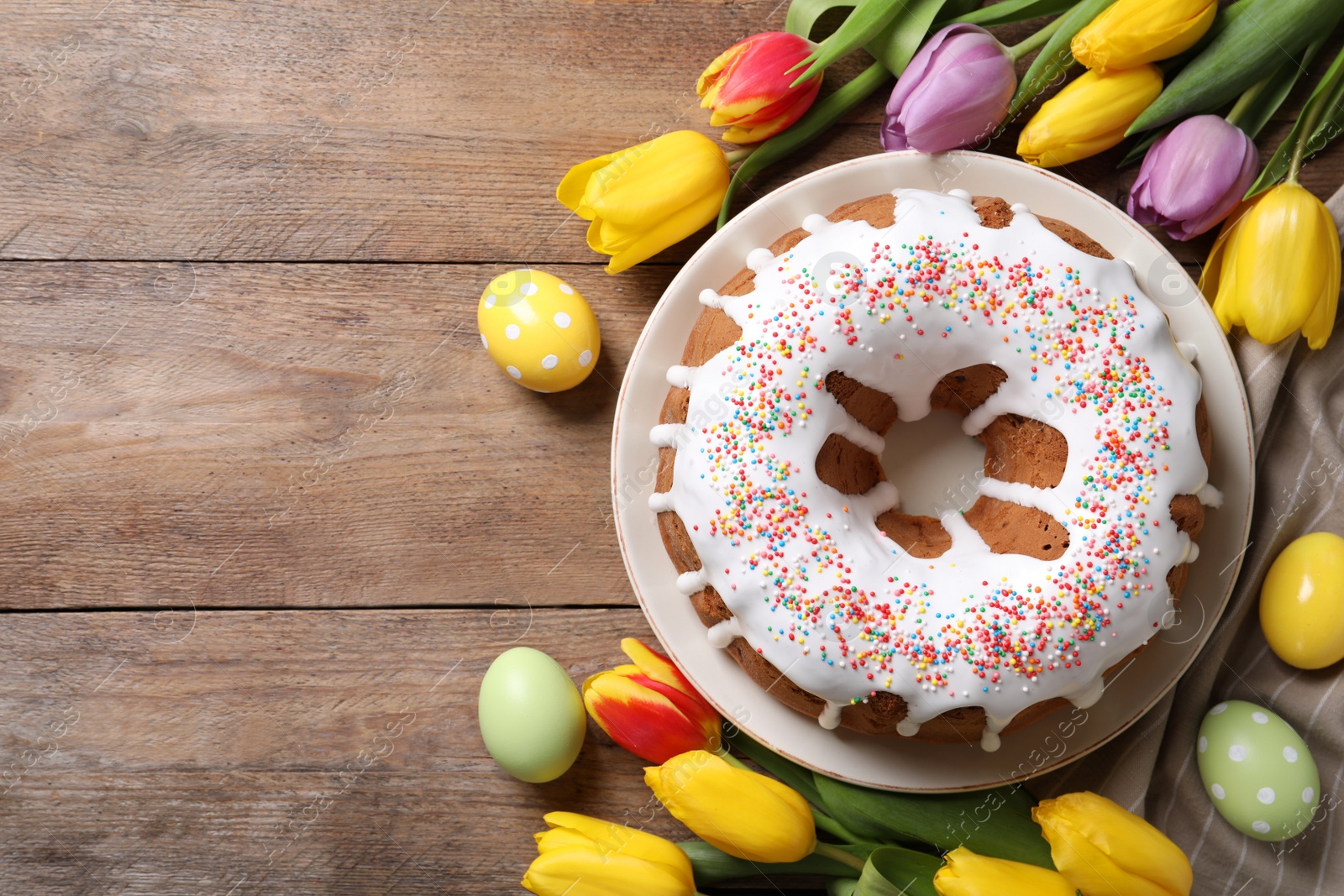 Photo of Glazed Easter cake with sprinkles, painted eggs and tulips on wooden table, flat lay. Space for text