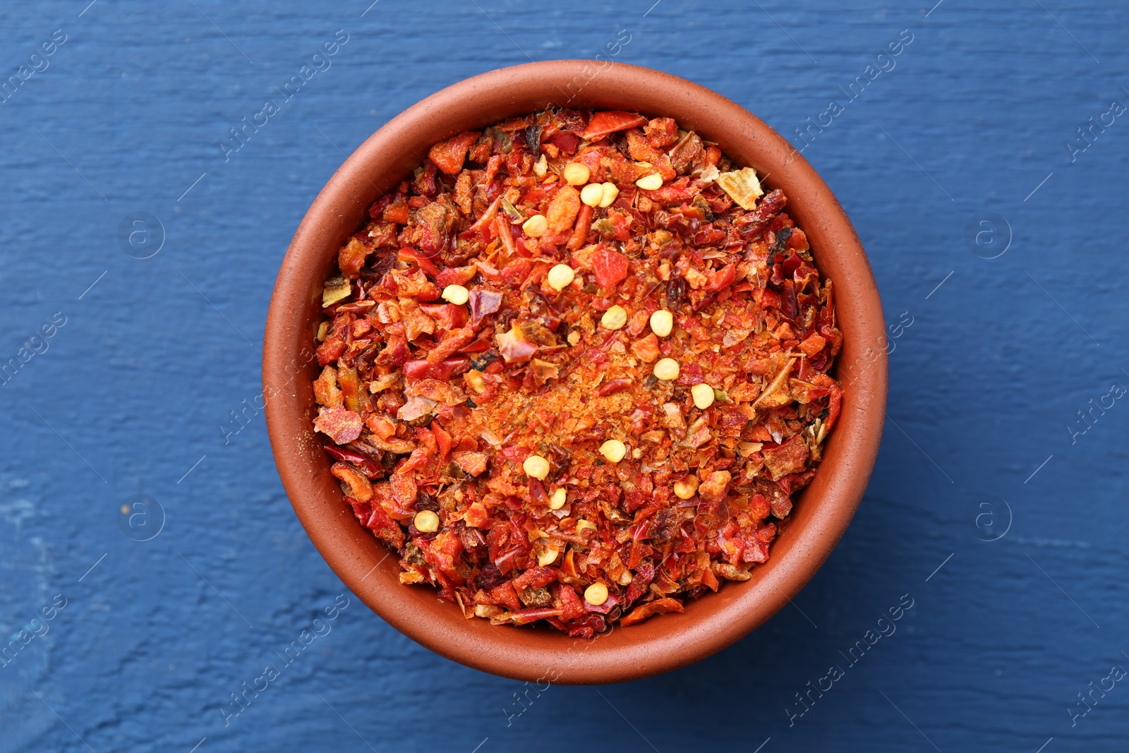 Photo of Chili pepper flakes in bowl on blue wooden table, top view