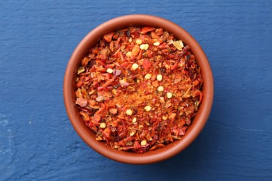 Photo of Chili pepper flakes in bowl on blue wooden table, top view