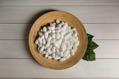 Photo of Silk cocoons with plate and mulberry leaves on white wooden table, flat lay