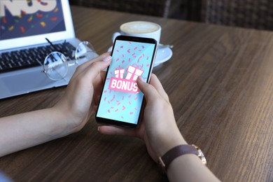Image of Bonus gaining. Woman using smartphone at wooden table, closeup. Illustration of open gift boxes, word and falling confetti on device screen