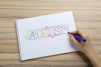 Photo of Woman writing word Autism inside jigsaw puzzle pieces in sketchbook at wooden table, top view