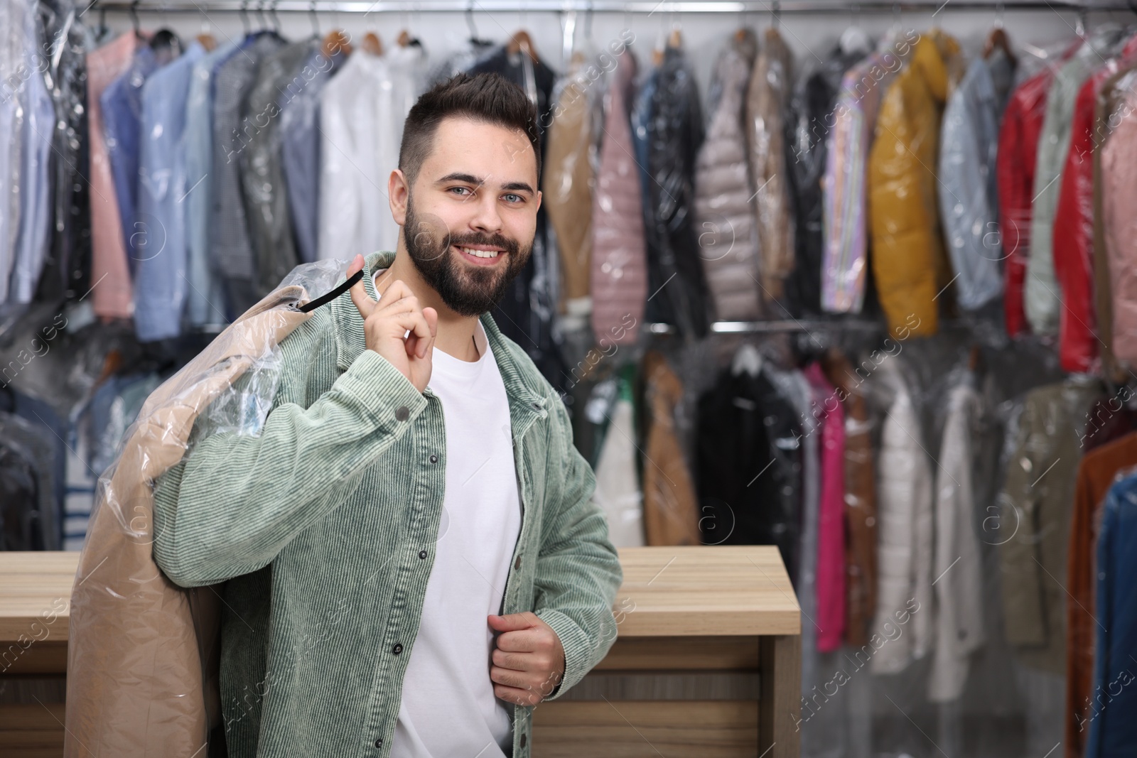 Photo of Dry-cleaning service. Happy man holding hanger with coat in plastic bag indoors, space for text