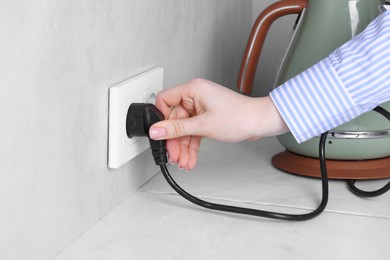 Photo of Woman plugging electric kettle into power socket at white table indoors, closeup