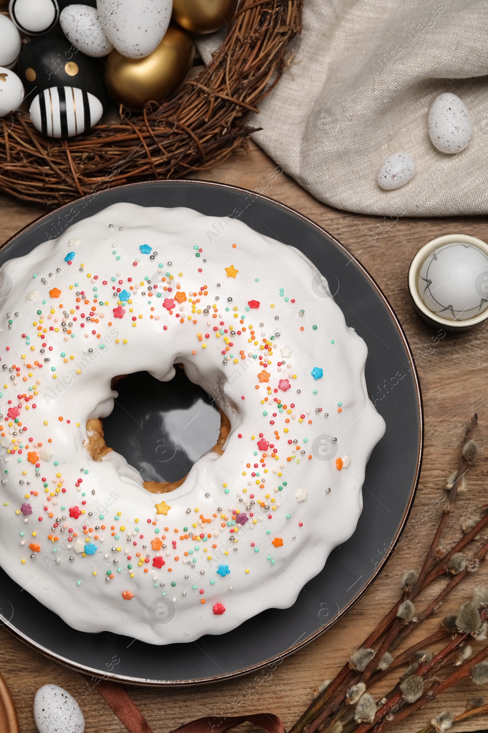 Photo of Easter cake with sprinkles, painted eggs and willow branches on wooden table, flat lay