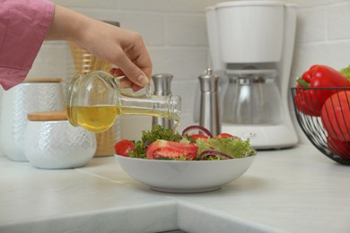 Woman pouring oil from jug into bowl with salad in kitchen, closeup