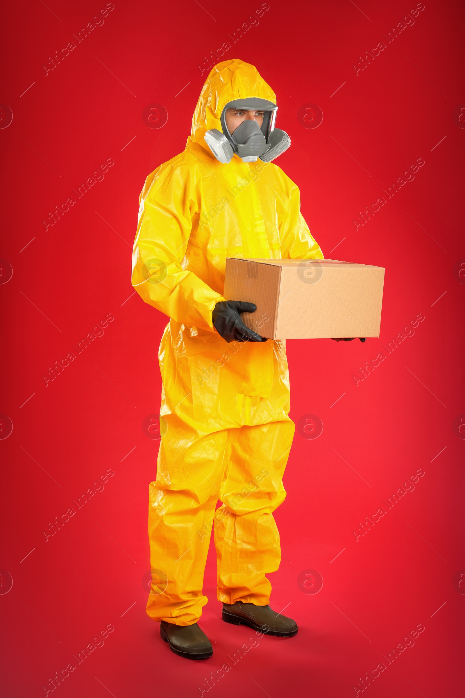 Photo of Man wearing chemical protective suit with cardboard box on red background. Prevention of virus spread