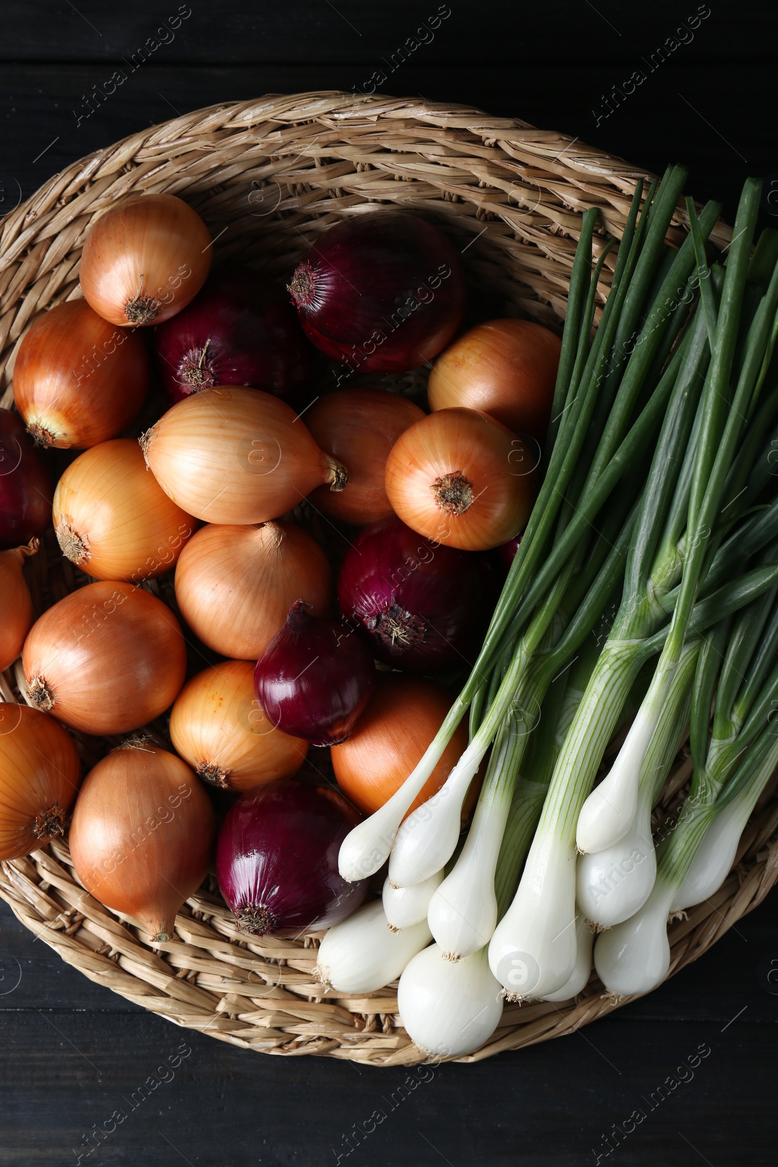 Photo of Wicker mat with different kinds of onions on black wooden table, top view