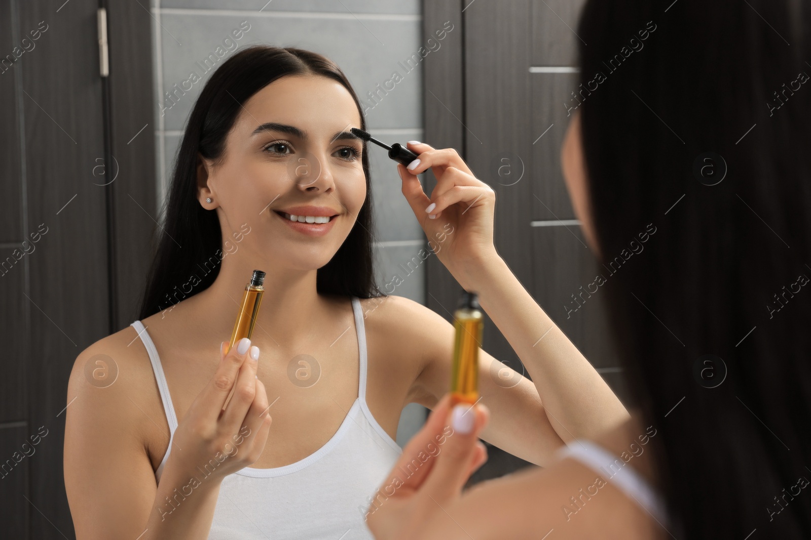 Photo of Young woman applying oil onto eyelashes near mirror indoors