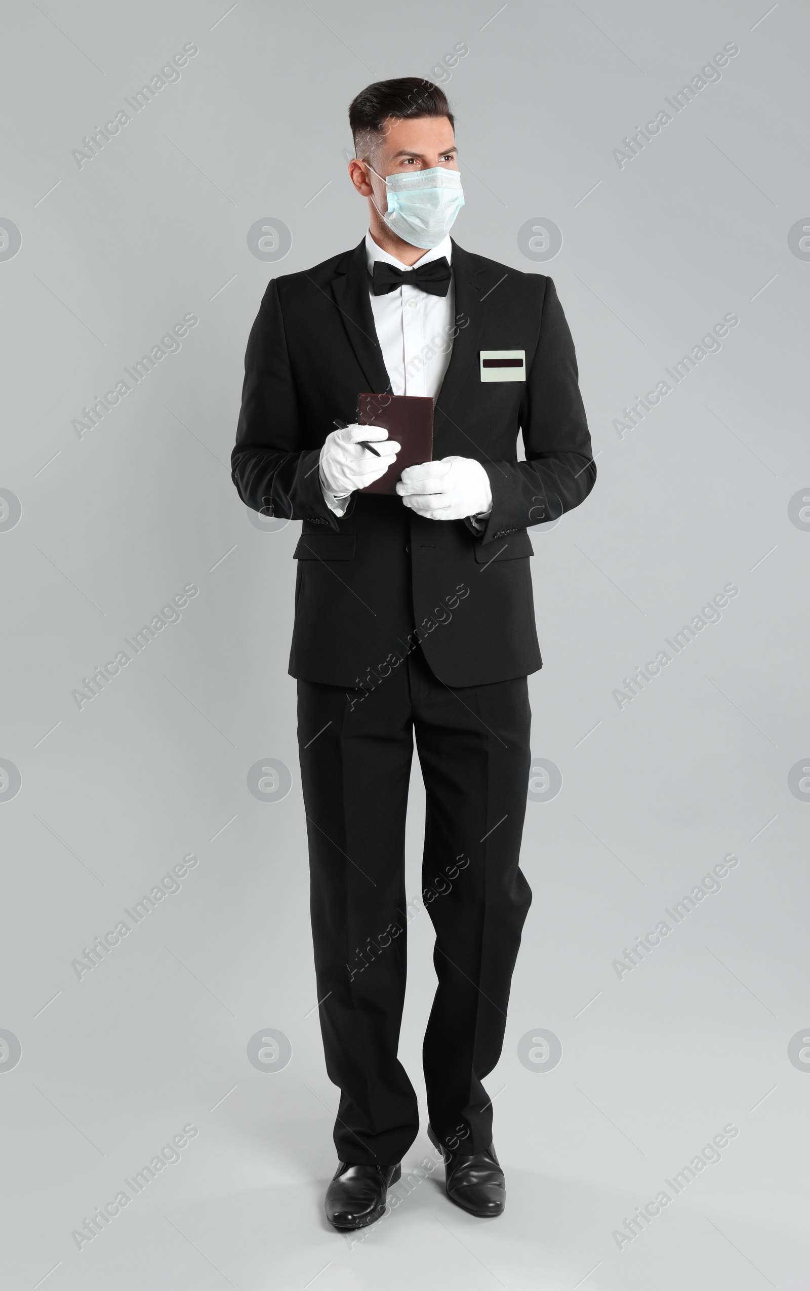 Photo of Waiter in medical face mask with notepad on light grey background