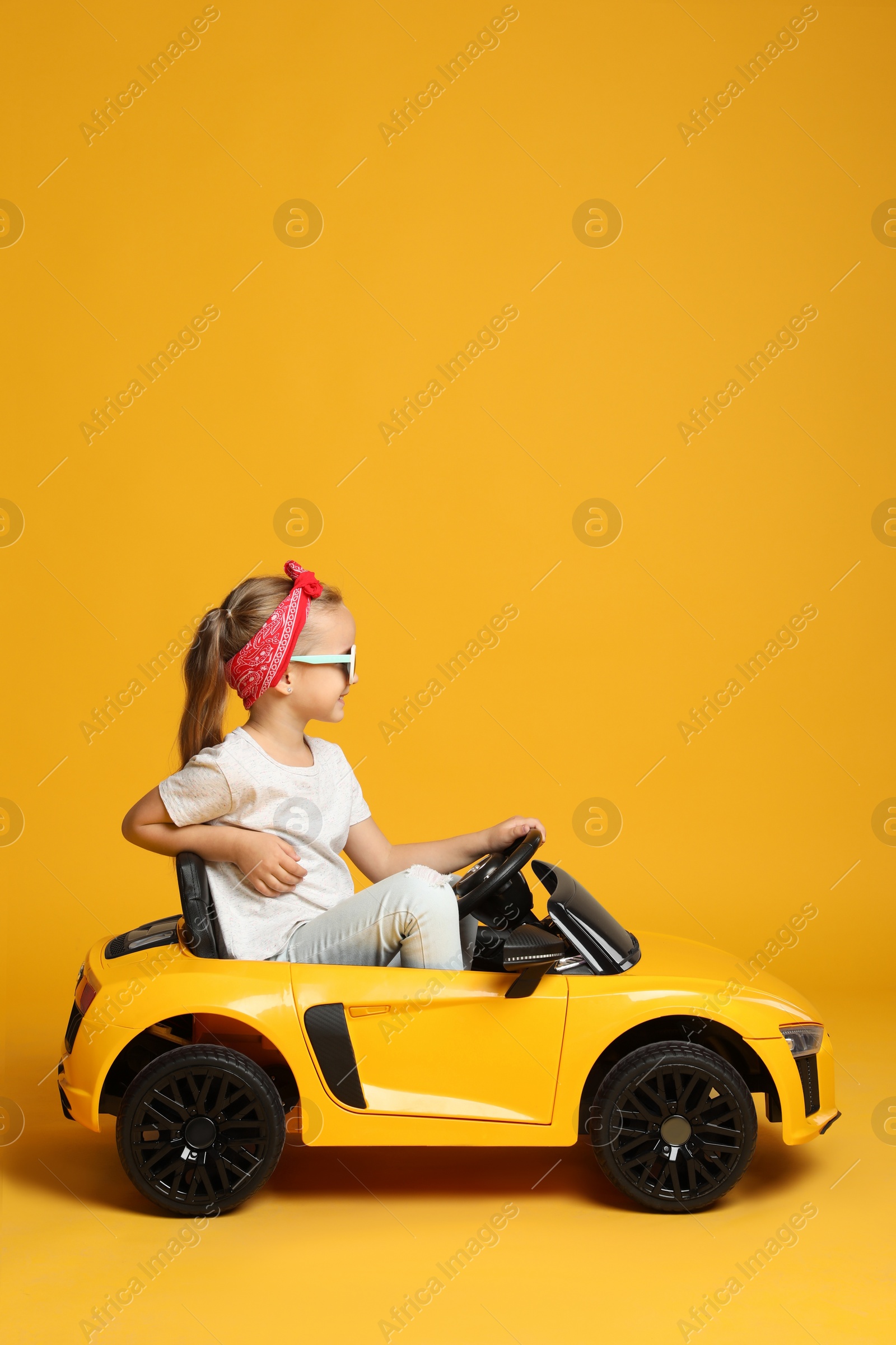 Photo of Cute little girl driving children's electric toy car on yellow background