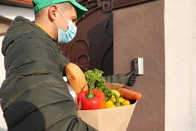 Courier in medical mask holding paper bag with groceries and ringing doorbell outdoors. Delivery service during quarantine due to Covid-19 outbreak