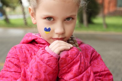 Photo of Little girl with drawing of Ukrainian flag on face in heart shape outdoors, closeup