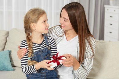 Photo of Cute little girl presenting her mother with gift on sofa at home