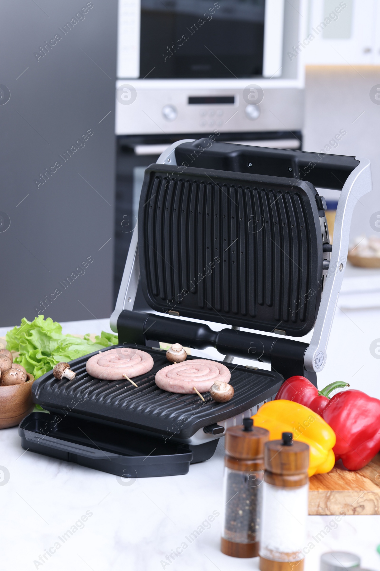 Photo of Electric grill with homemade sausages, mushrooms, vegetables and spices on white table in kitchen