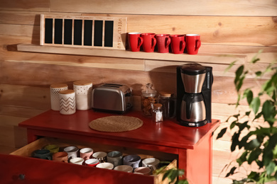 Photo of Open drawer with cups and coffeemaker near wooden wall indoors